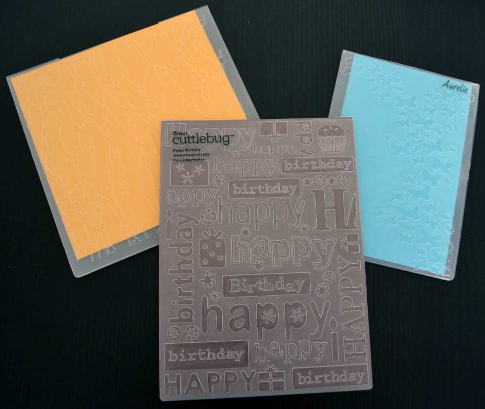 Drei Beispiele für Prägefolder: links ein weihnachtliches Motiv von Marianne Design (Celestial), rechts ein Schmetterlingsmotiv von Aurelie (Butterfly Dreams), vorne mittig ein Geburtstagshintergrund von CriCut (Cuttlebug: Happy Birthday).